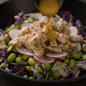 a salad with chicken and edamame soy beans with dressing being poured on top with a spoon